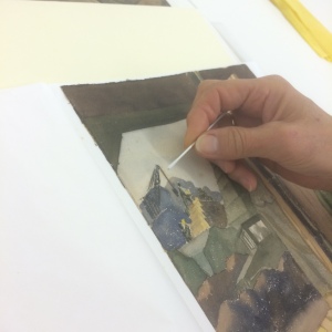 In this image, paper conservator Karen Zukor is examining and testing a watercolor painting from 1943 Adak Island by Warren Beach.  The artwork has some severe spotty staining on the back that is beginning to show through to the front.  A later to posting will detail some of our strategies to control light levels in the upcoming exhibits.